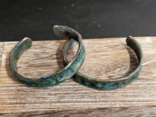 Load image into Gallery viewer, Two handmade open cuff bracelets with a textured, rustic finish in dark green and bronze tones, displayed on a wooden surface.