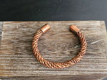 Load image into Gallery viewer, A handmade copper-toned twisted wire cuff bracelet with cylindrical end caps, displayed on a rustic wooden board against a dark background.