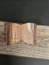 Load image into Gallery viewer, A copper-colored metal cuff bracelet with intricate floral engravings, resting on a weathered wooden surface against a dark background.