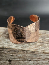 Load image into Gallery viewer, A copper-colored metal cuff bracelet with intricate floral engravings, resting on a rustic wooden surface against a dark gray background.