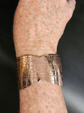 Load image into Gallery viewer, A close-up of a person's forearm wearing a wide, ornate metallic cuff bracelet with engraved floral patterns and a wavy cut edge, set against a dark background.