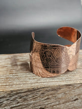 Load image into Gallery viewer, A copper-colored metal cuff bracelet with intricate floral engravings, resting on a rustic wooden surface.