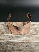 Load image into Gallery viewer, A hammered copper cuff bracelet with engraved floral patterns and a fringed edge, displayed on a rustic wooden surface against a dark background.