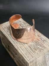 Load image into Gallery viewer, A hammered copper cuff bracelet with engraved floral patterns, resting on a rustic wooden block against a dark background.
