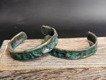 Load image into Gallery viewer, Two textured open cuff bracelets with a distressed green and bronze patina, displayed on a rustic wooden surface against a dark background.
