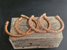 Load image into Gallery viewer, Four copper-toned twisted wire cuffs or rings displayed on a rustic wooden block against a dark background.