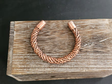 Load image into Gallery viewer, A copper-colored twisted rope cuff bracelet with metal end caps, displayed on a rustic wooden block against a dark background.