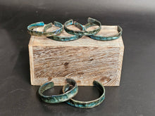 Load image into Gallery viewer, Three open cuff bracelets with a distressed turquoise and dark metallic finish, displayed on and around a rustic wooden block against a dark gray background.